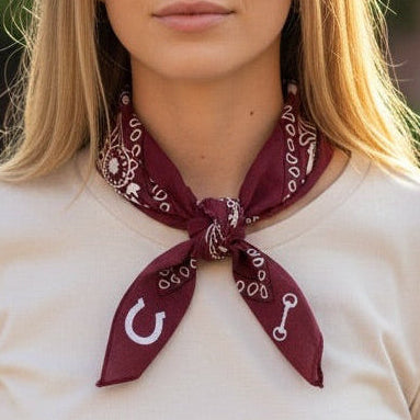 A woman wearing a cream-colored t-shirt and blue jeans, accessorized with a maroon and white patterned bandana tied around her neck, set against a blurred outdoor background.