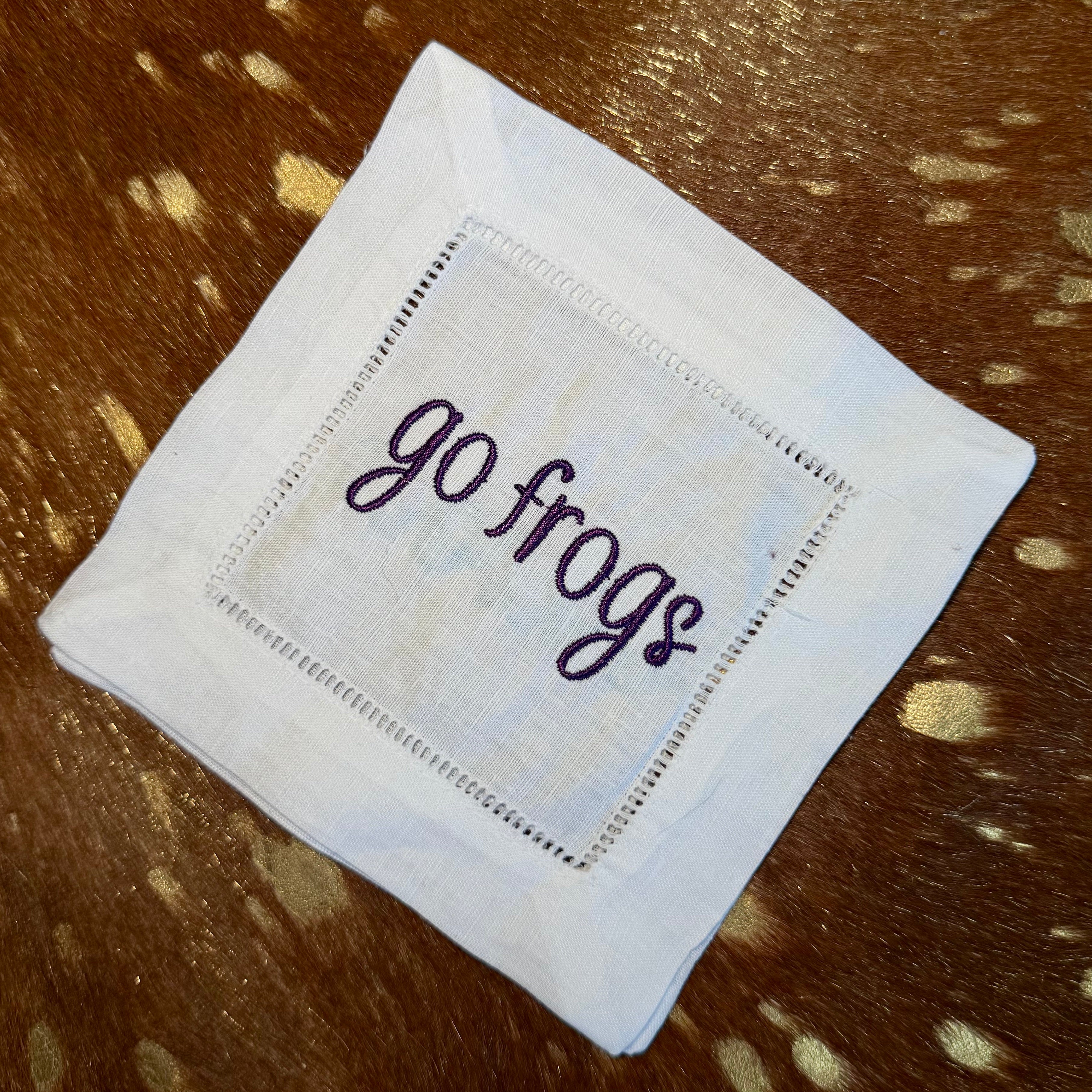 White linen-cotton cocktail napkin with purple Go Frogs embroidery styled on cowhide